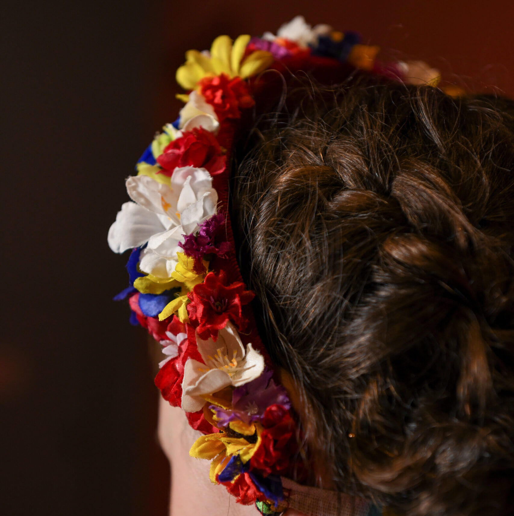 Shot from the back, a dancer wears a headband made of white red and yellow flowers with colorful ribbons hanging from the bottom.