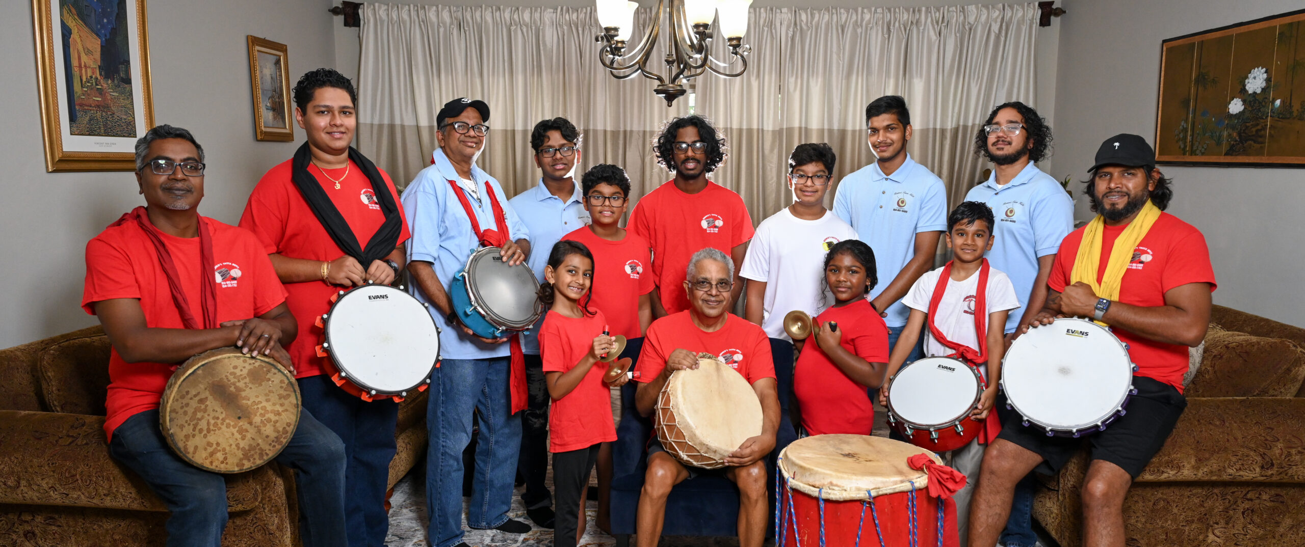 A large ensemble of various ages stands around Romeo Ragbir, seated in the middle. Some members have drums hanging from their necks, and two younger players hold hand cymbals.