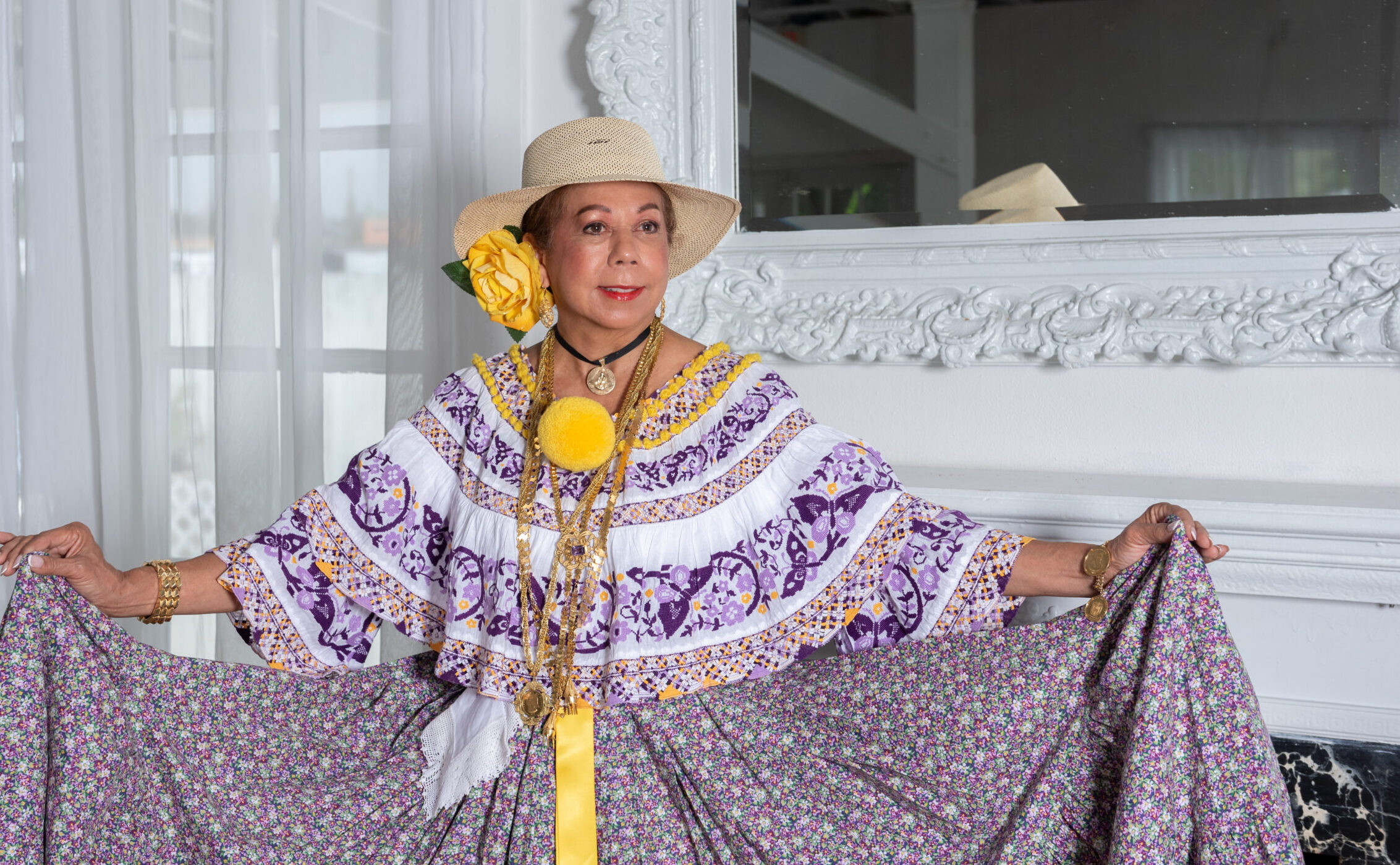 A woman in a purple and white embroidered dress and a straw hat poses with he skirt stretched out to the sides.
