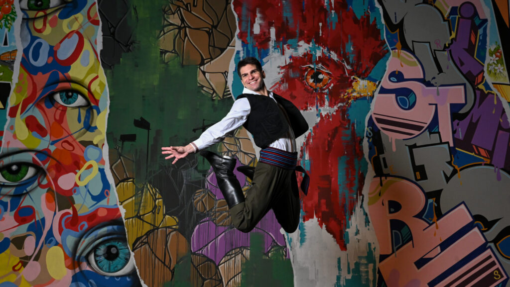 A dancer jumps in the air with his legs and arms stretched behind him. He is in front of a colorful backdrop and is wearing a white shirt with embroidered designs, a black vest, green pants, and a colorful belt