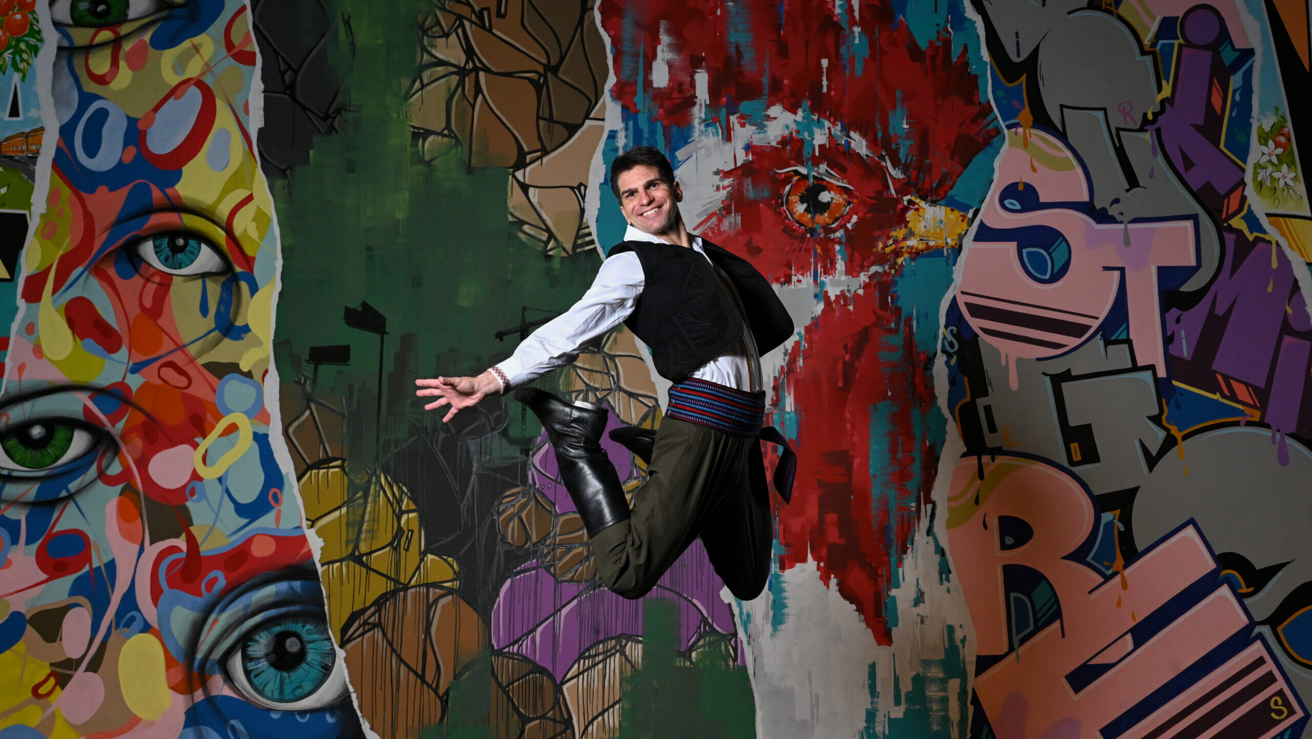 A dancer jumps in the air with his legs and arms stretched behind him. He is in front of a colorful backdrop and is wearing a white shirt with embroidered designs, a black vest, green pants, and a colorful belt