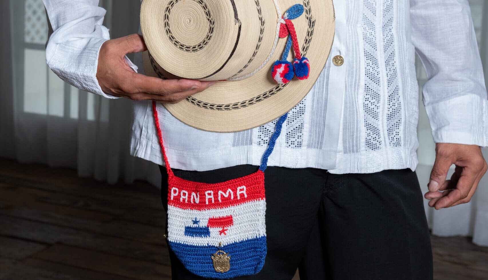 A man holds a straw hat held to his side. The hat has embroidery details in blue and red. He also wears a small emboridered purse that says the word 