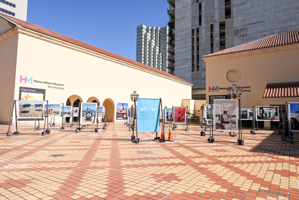 View of HistoryMiami Museum from the Cultural Plaza. Two beige buildings sit on an orange and yellow tiled plaza. A photo exhibit displayed on wooden frames sit between them. In the distance, a building is being constructed against a blue sky.