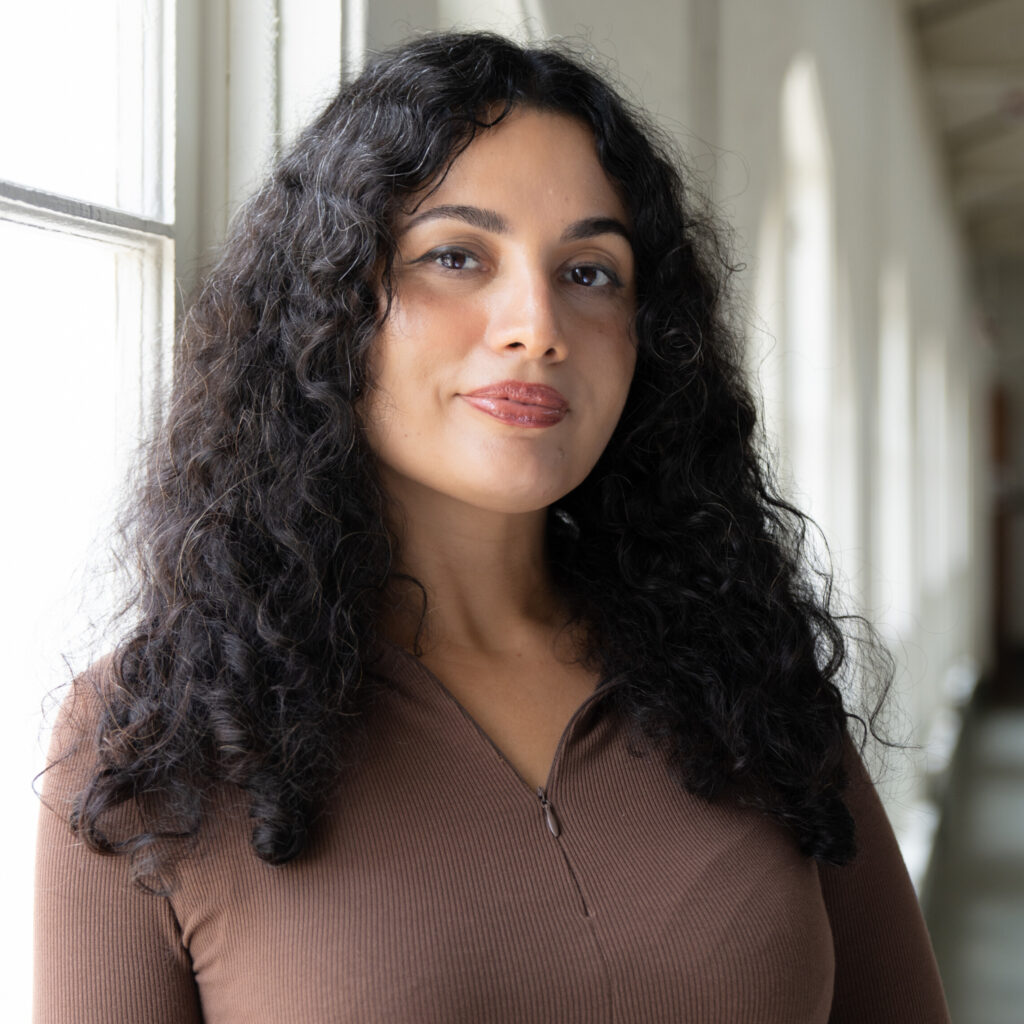 A person with dark brown curly hair looks towards the camera.