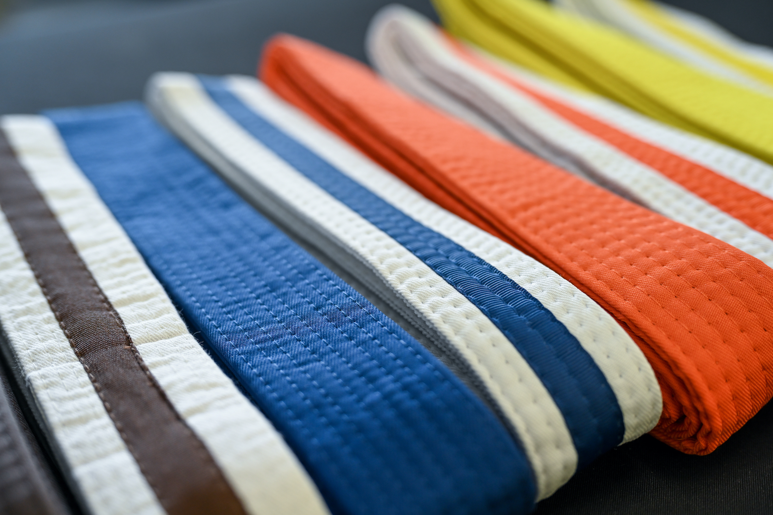 Taekwondo belts of various colors lie on a table in a row. Some are white with a color stripe and some are solid colors.