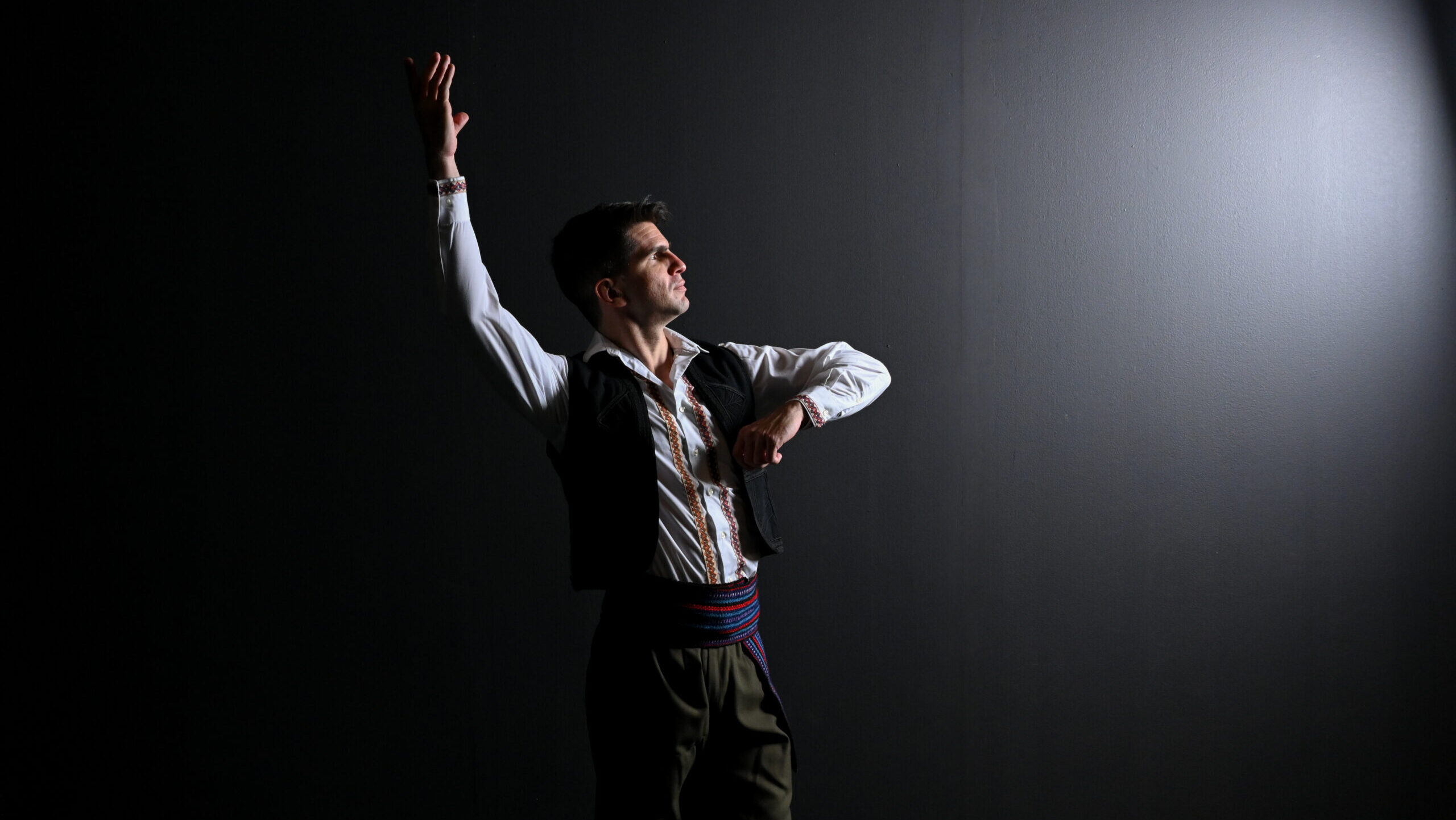 A man stands against a black background with a white light shining on him. He is looking at the light and standing with one arm in the air and one bent at the elbow