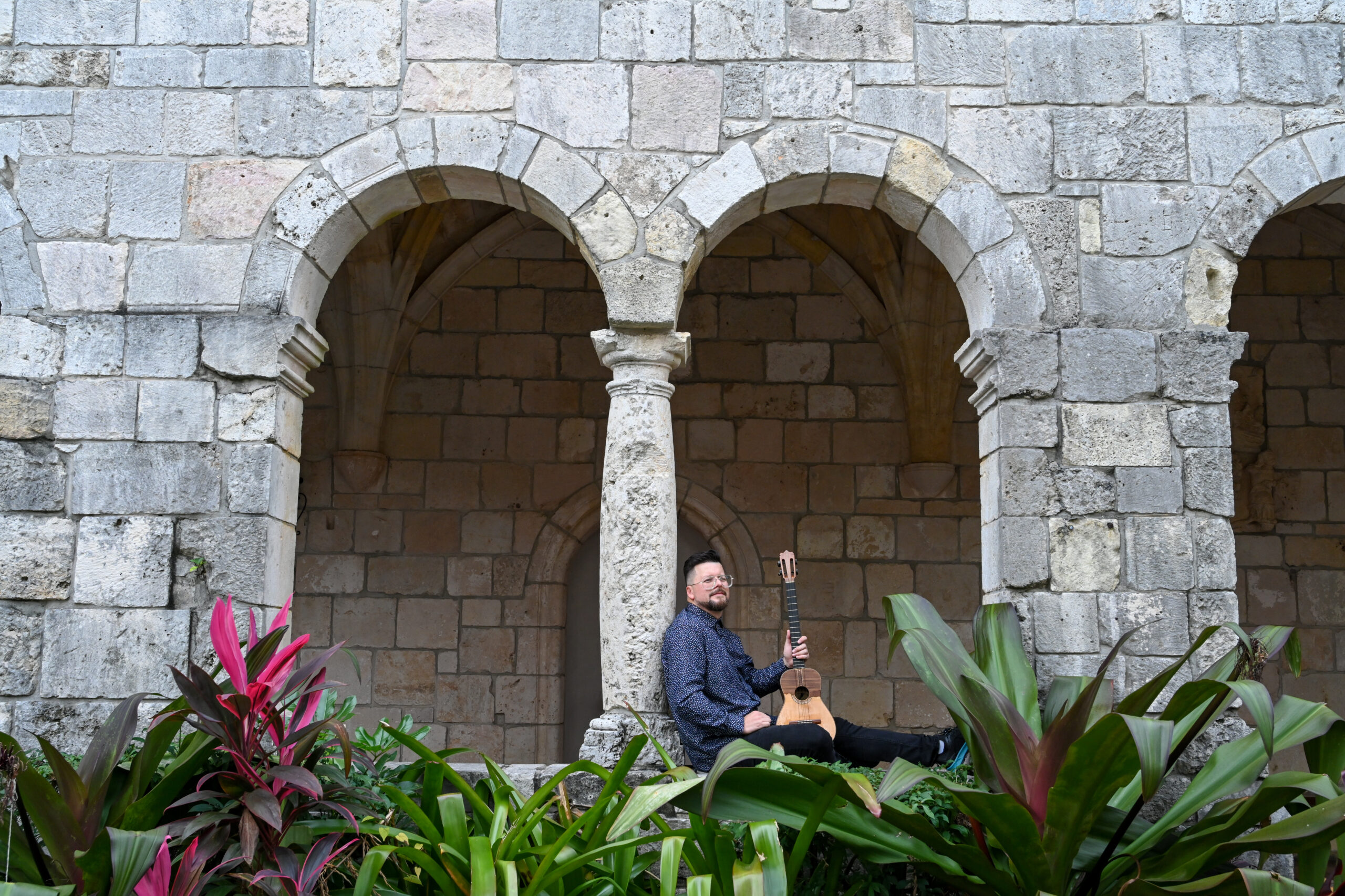 Henry is sitting under two arches, leaning on the pillar between them while holding his cuatro guitar upright in his hands.