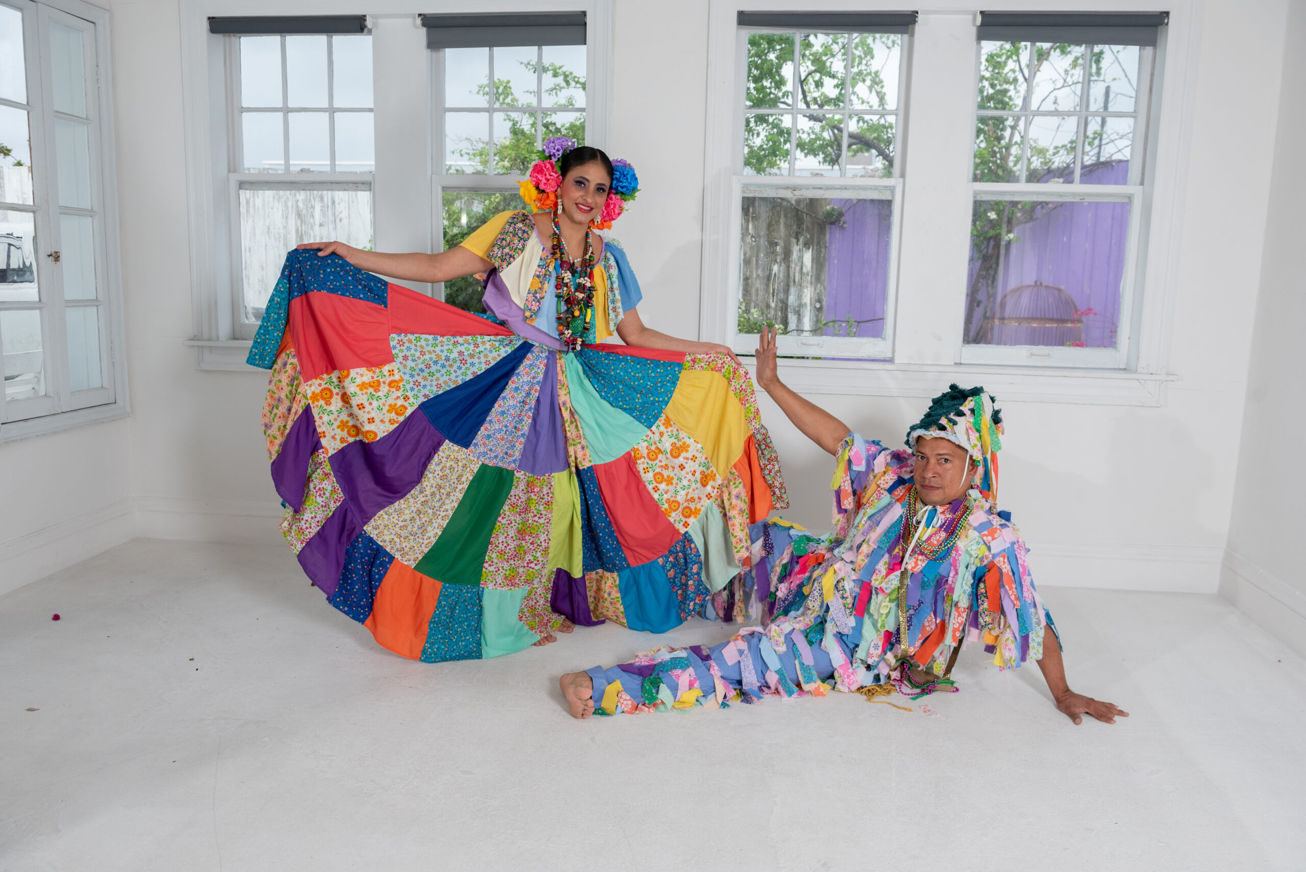 Two dancers in very colorful costumes pose. The woman holds out her colorful skirt to the sides. The man lays on the floor in front of her with one arm stretched out towards her. he wears an outfit with colorful strips of cloth hanging and a tall hat with strips of colorful cloth.