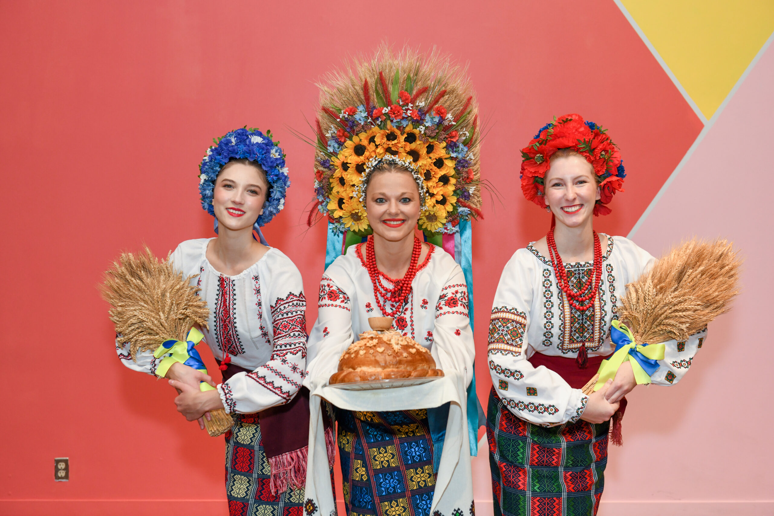 Three dancers pose. The one in the middle holds a bread offering and the two on the sides hold bundles of wheat.