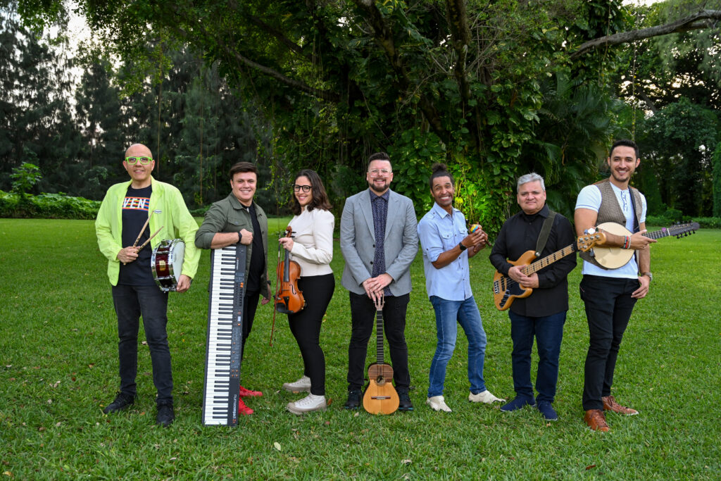 The Henry Linarez Ensemble stand in the grass, each holding their instrument: snare drum, keyboard, violin, cuatro, maracas, bass, mandolin