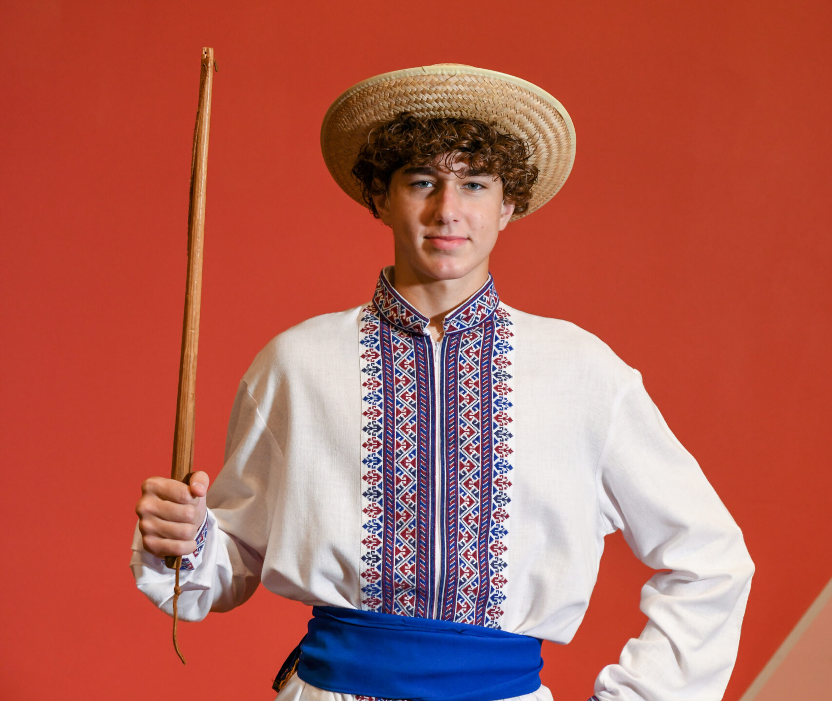 a young male dancer wears a straw hat, a blue embroidered shirt, and a blue sash.