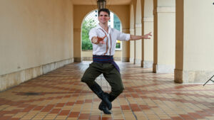 A dancer poses with his toes pointed towards the center and his arms outstretched. He outdoors in a corridor with hanging lamps and is wearing a white shirt with embroidered designs, green pants, and a colorful belt
