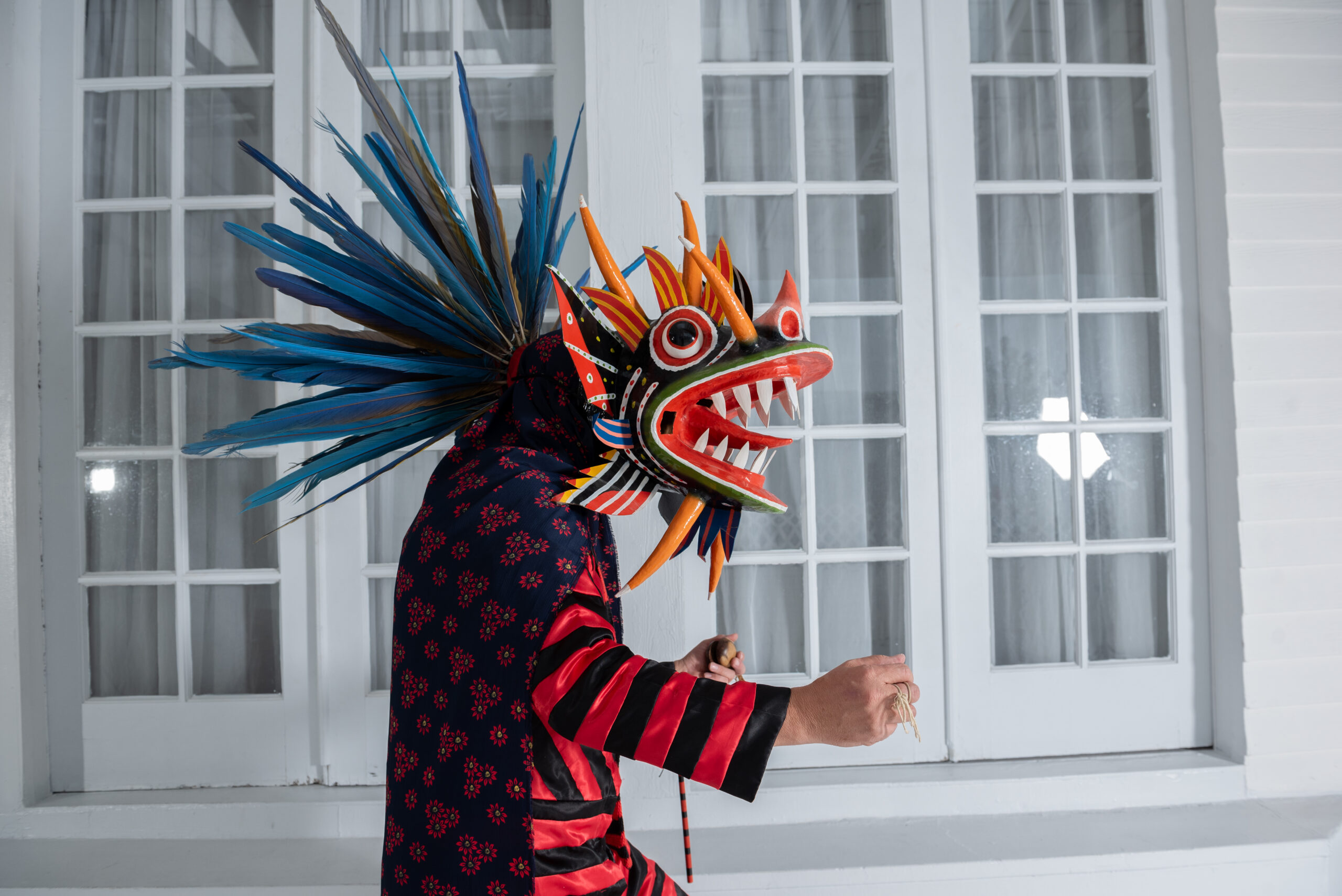 A dancer wears a red and black striped shirt and cape. He wears a colorful mask depicting a devil with large teeth, horns, and with feathers fanning out on top of the head.