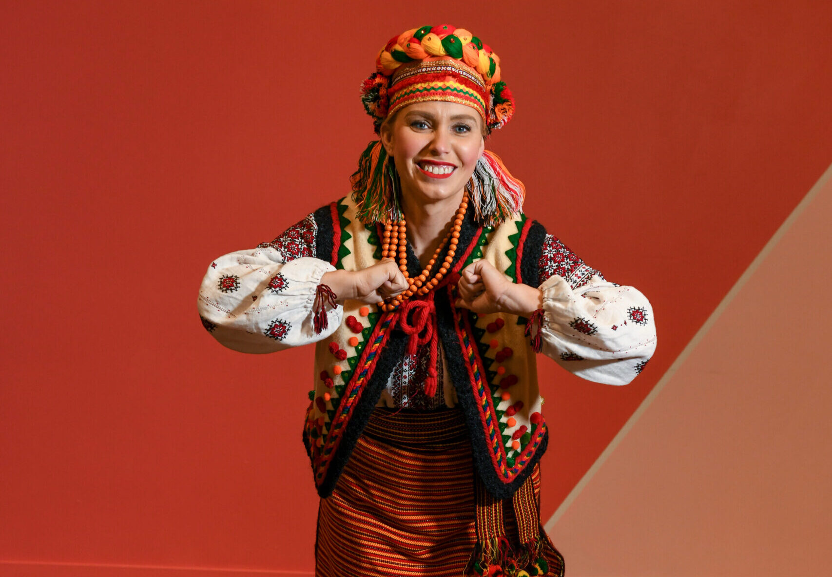 A dancer wears a green, orange, and yellow hat with an embroidered vest and long skirt. She is holding out her elbows with her thumbs pointed into her chest while leaning forward and smiling.