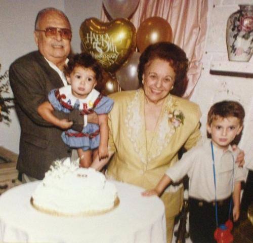 An older man holds a small girl next to an older woman with her arm around a boy. They're standing in front of a cake with balloons behind them.