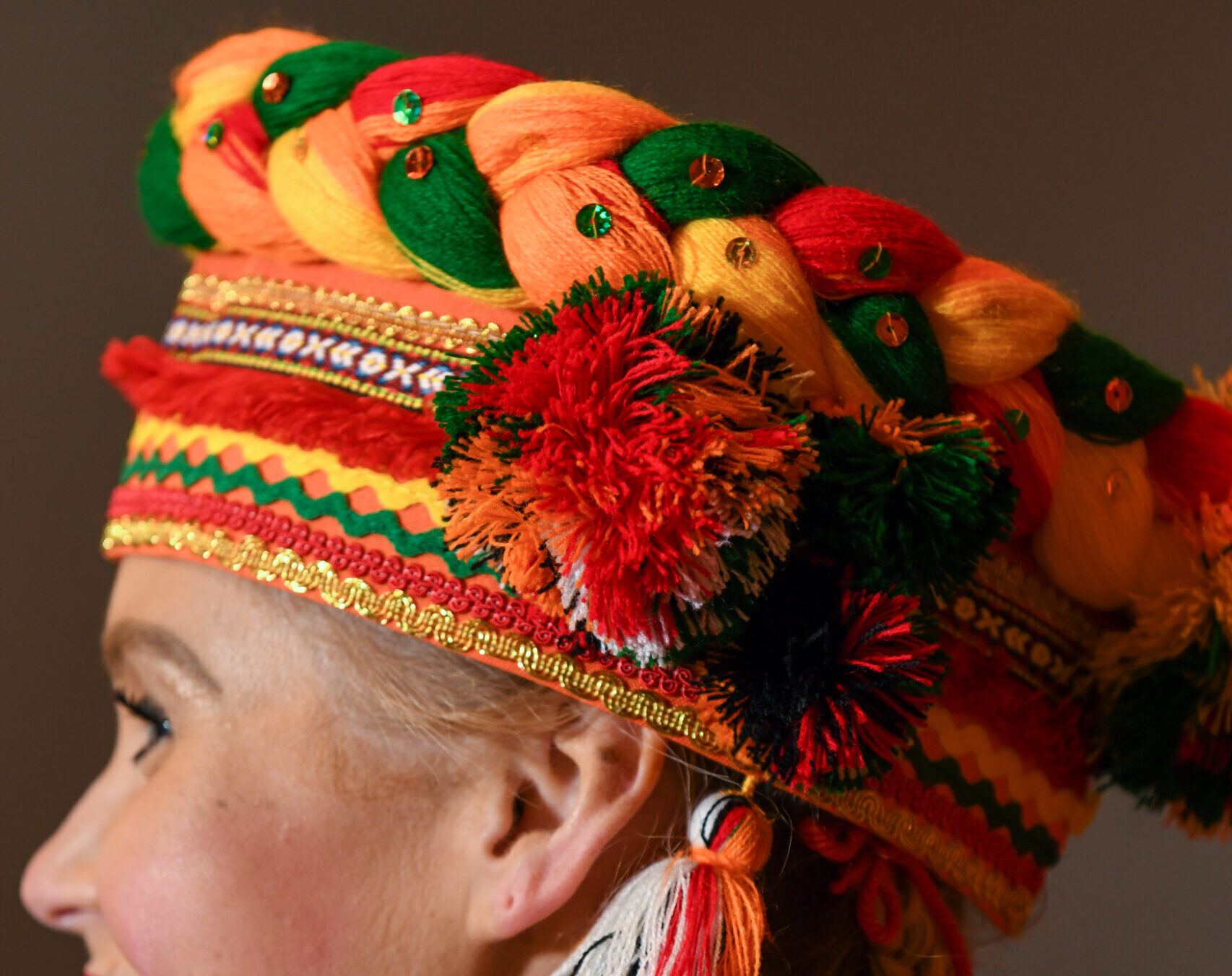A hat made of bright orange, yellow, green, and red wool. Thick strands of wool are braided to make the top of the hat with rows of ribbon forming the piece around the forehead.