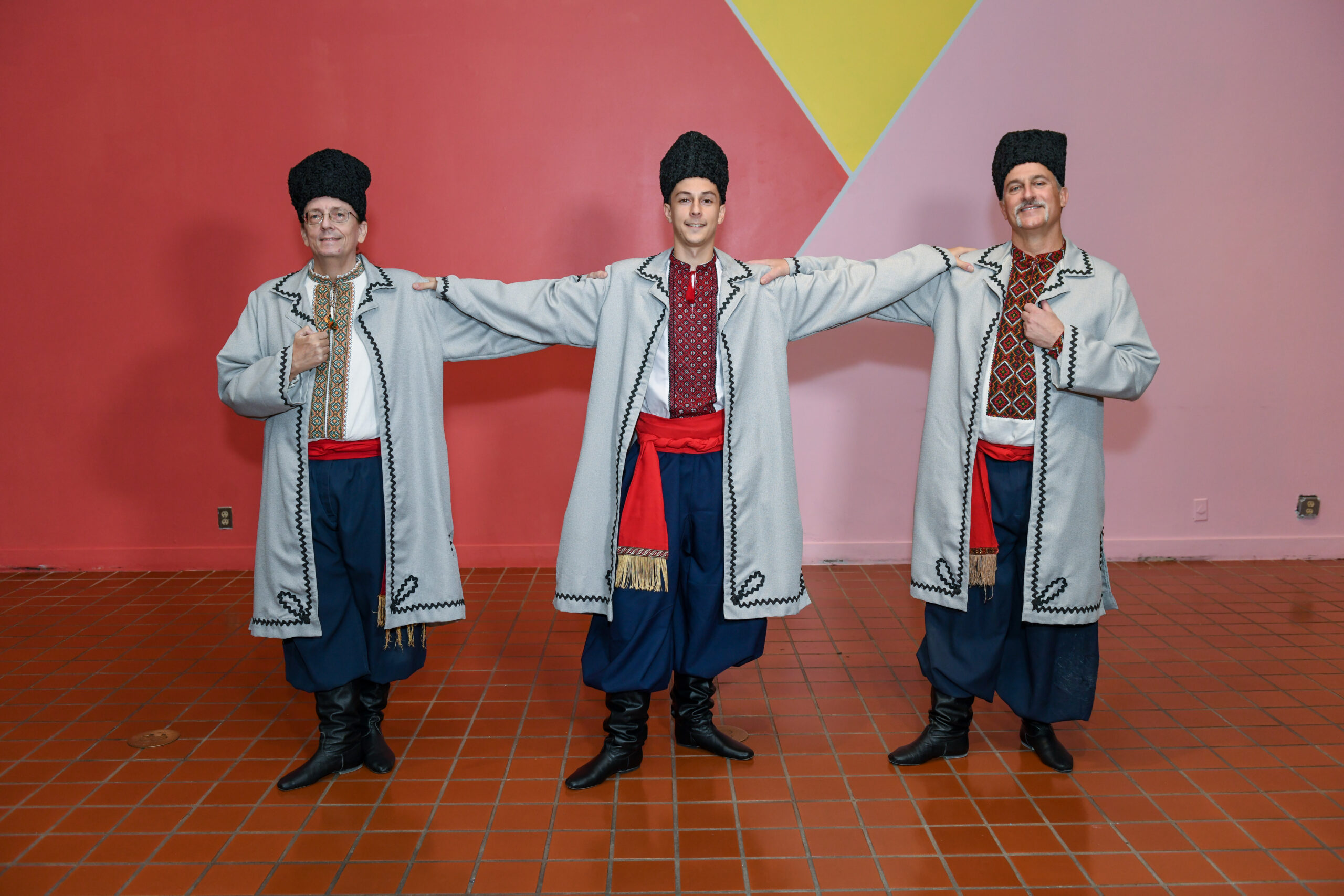 Three men pose with their arms outstretched on each others shoulders. they wear black tall hats, grey coats, embroidered shirts, and red sashes.