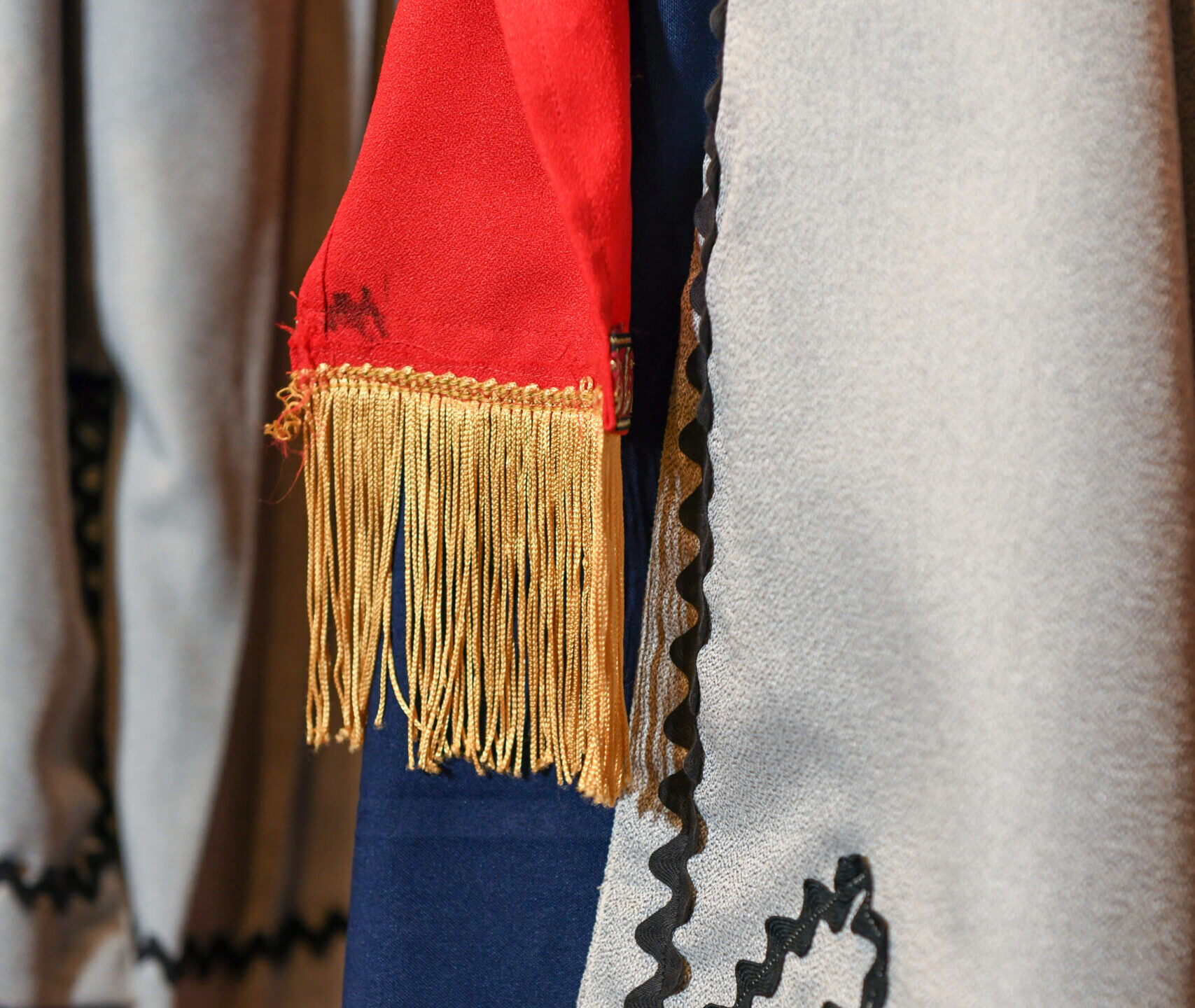 Detail of a grey coat with black embroidery and a red sash with yellow tassles.