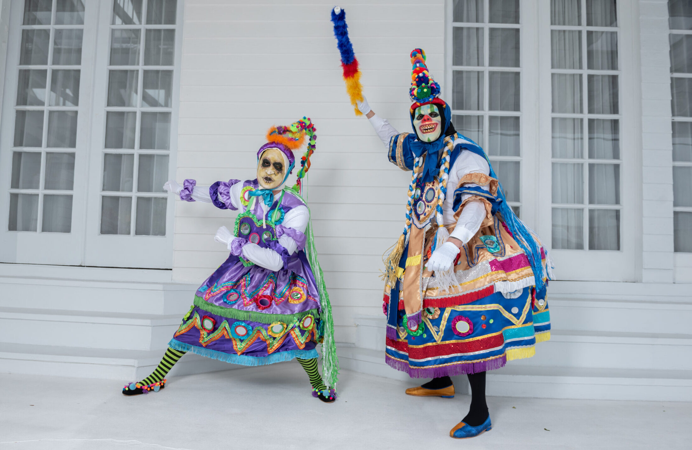 Two dancers in colorful tops and hooped skirts adorned in small mirrors pose for a photo. They have masks resembling clowns and elaborate headpieces with a colorful tail protruding from the tops of their heads.
