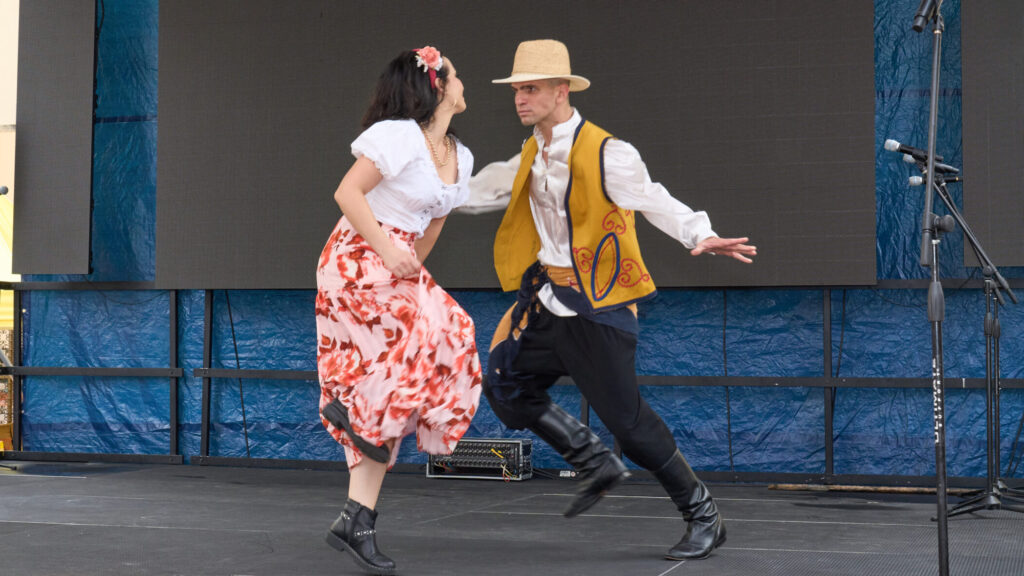 A couple dances on a stage. The male dancer advances towards the female dancer with his arms spread at his sides. The female dancer kicks one leg up as she dances towards him, holding her skirt in one hand.