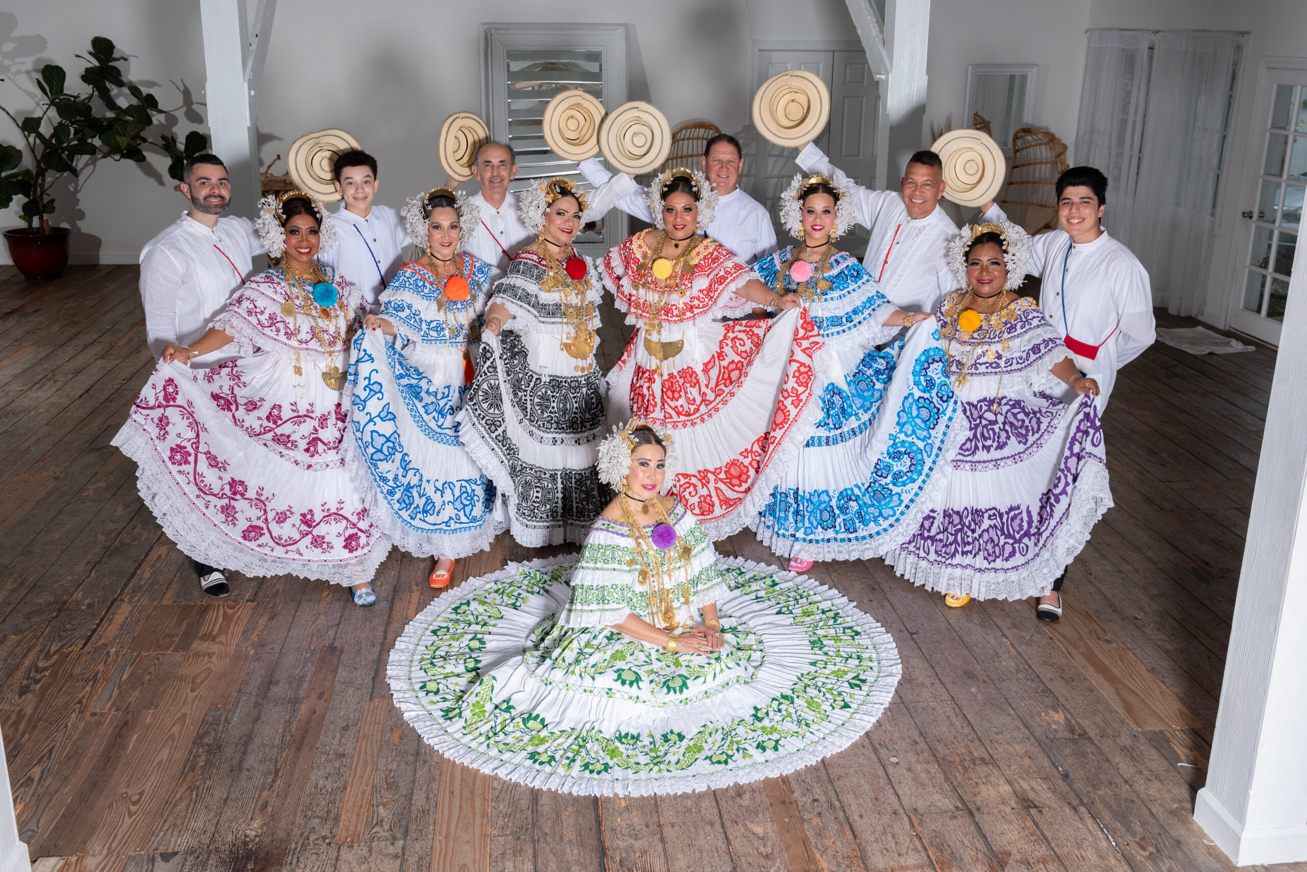 Seven women and six men pose for a photo. The women wear elaborate embroidered dresses in different colors with beaded headpieces and gold chains. The men stand behind the women and wear white shirts and hold up a straw hat in one hand. One woman sits in the front of the group with her skirt spread in a circle around her.