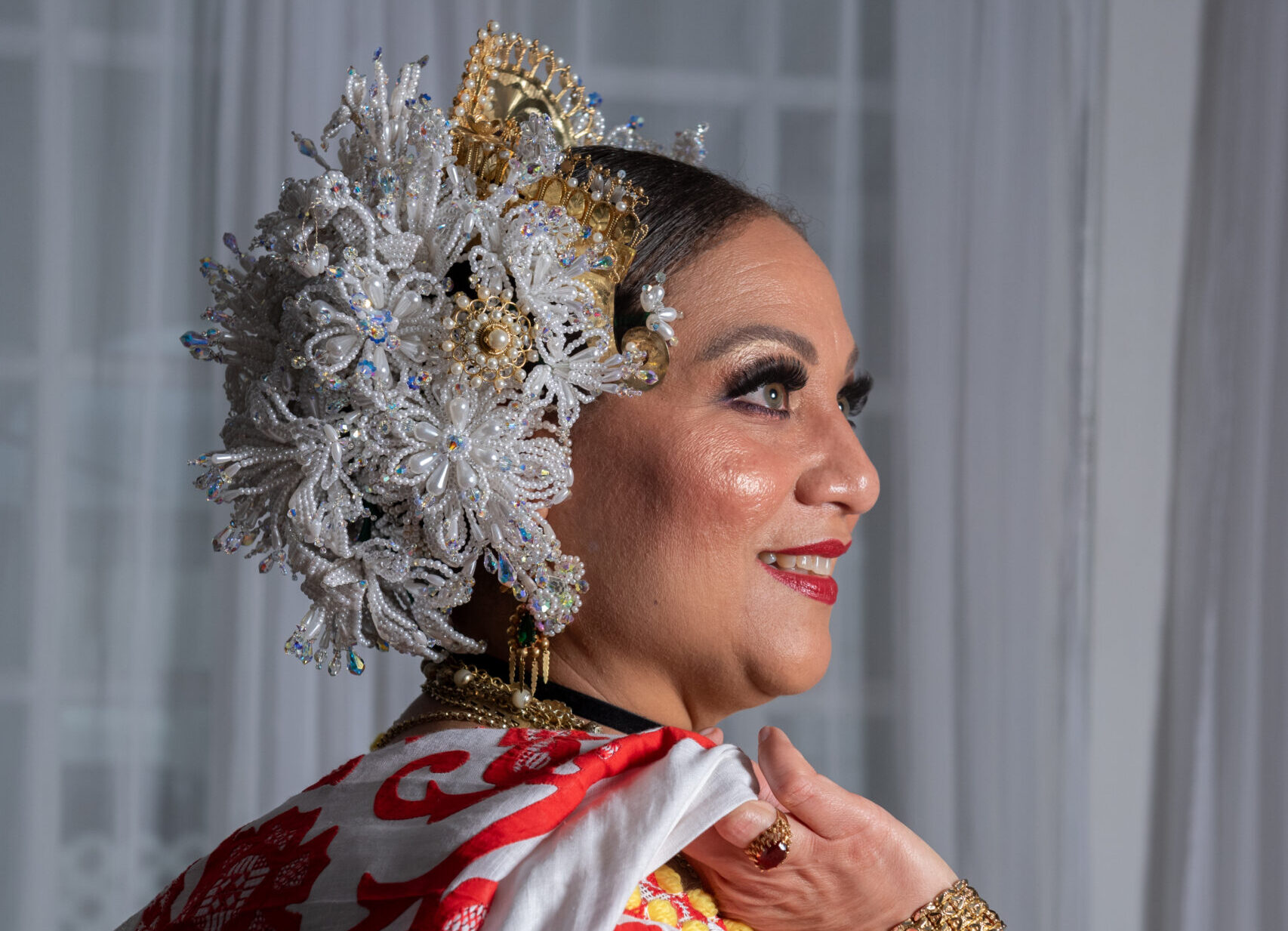 A woman in a red and white embroidered dress looks off to the side, showing the elaborate white and gold beaded headpiece on the side of her head.