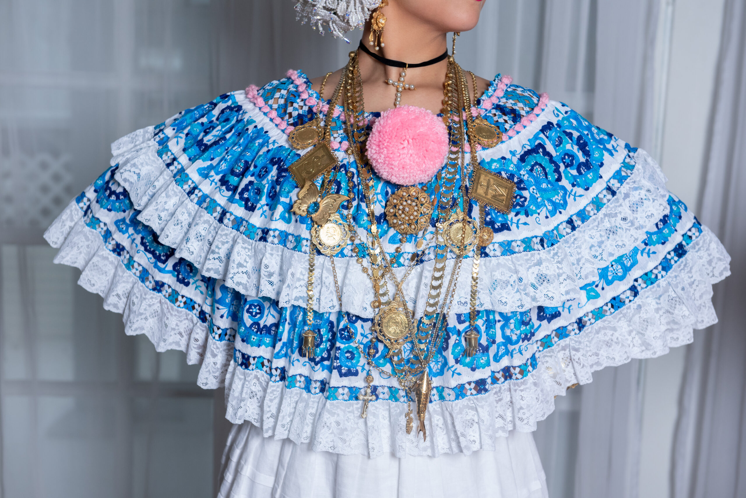 A close up shot of a woman in a blue and white embroidered blouse with pink accents. She wears various gold chains.