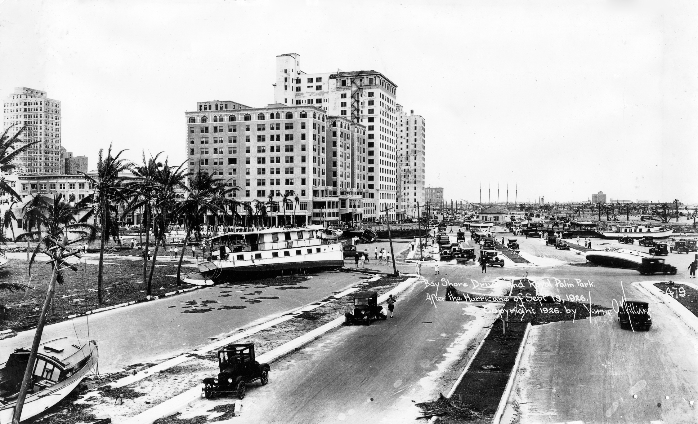 A black and white image of a street view in downtown Miami in the aftermath of a hurricane. Boats can be seen strewn along the street next to the bay with downed poles nearby.