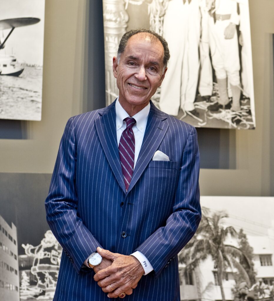 Dr. George, wearing a blue pinstripe suit, burgundy striped tie, and white shirt stands in front of a gallery of black and white archival images against a gray wall.