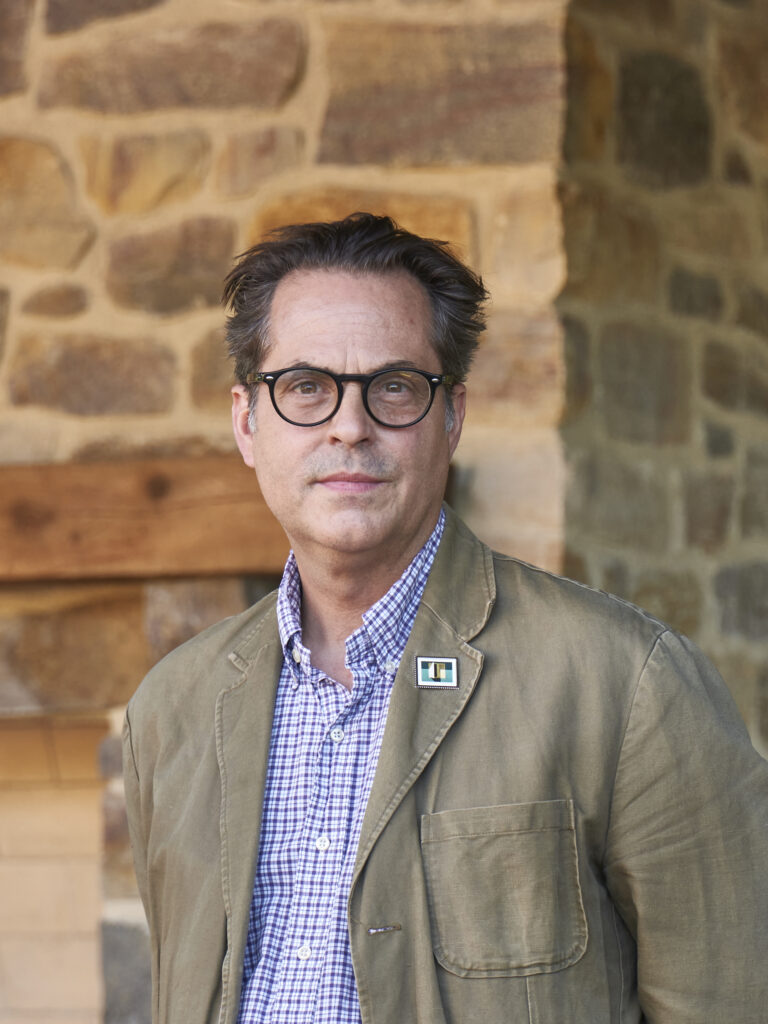 A person with short hair and dark rimmed glasses looks towards the camera. They wear a beige linen blazer and a gingham button down shirt.