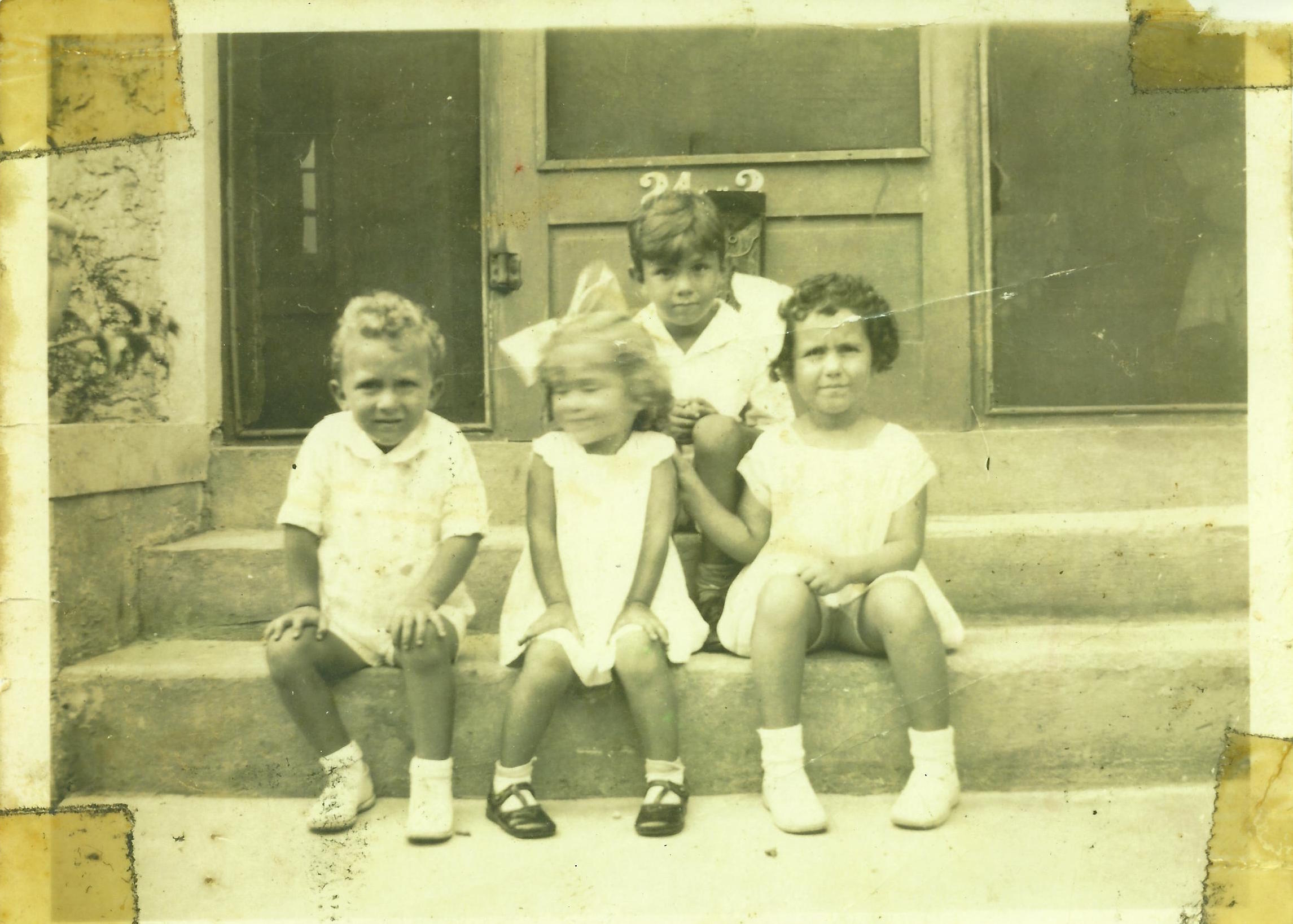 A black and white photo of four small children sitting on the steps of a building, three girls and one boy.