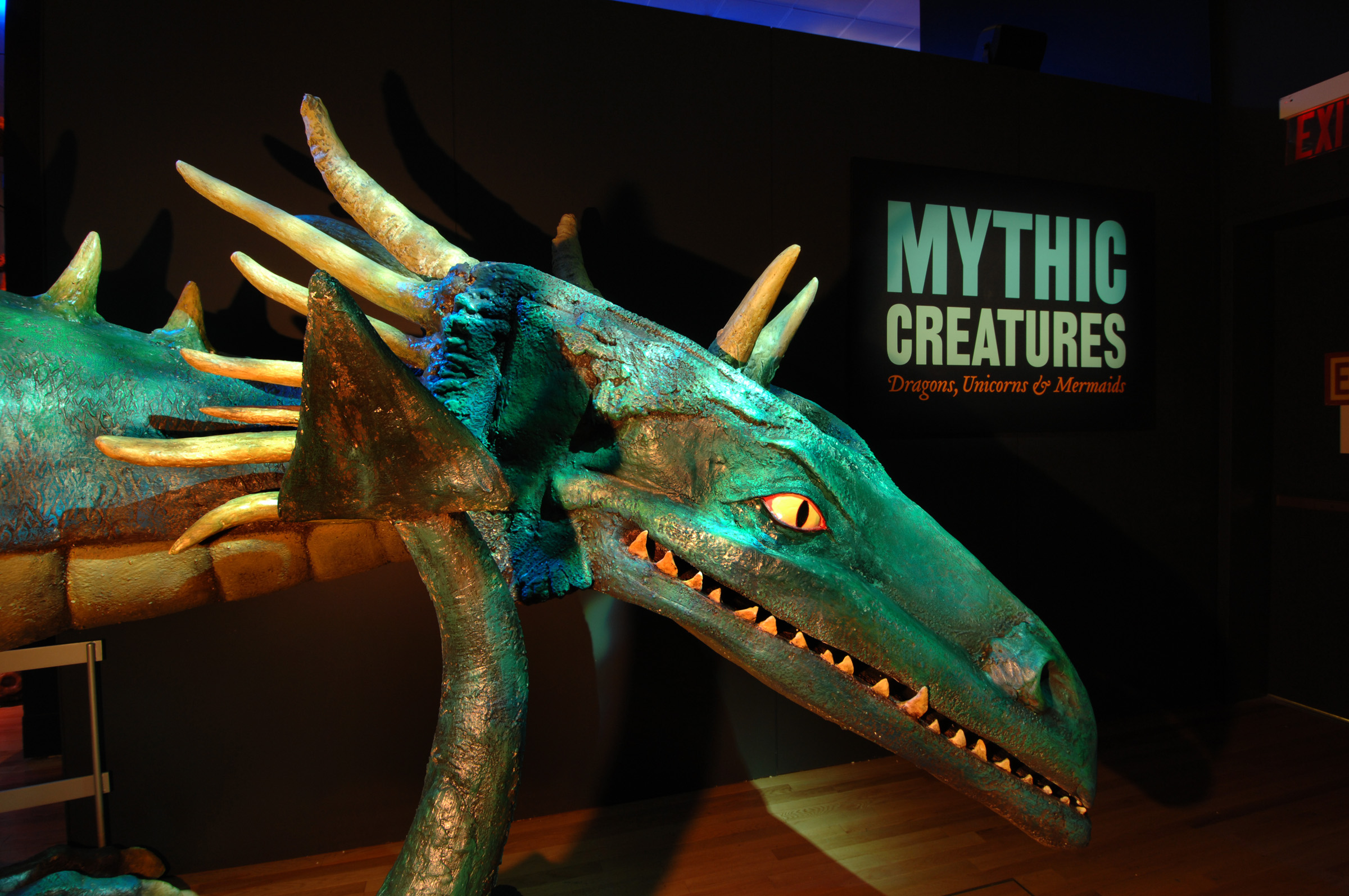 View of a dark exhibition with a large greenish blue model off a dragon that stands inside. On a dark wall, a sign reads, "Mythic Creatures."