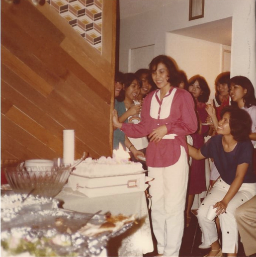 A woman stands smiling behind a cake with a group of woman behind her smiling at her