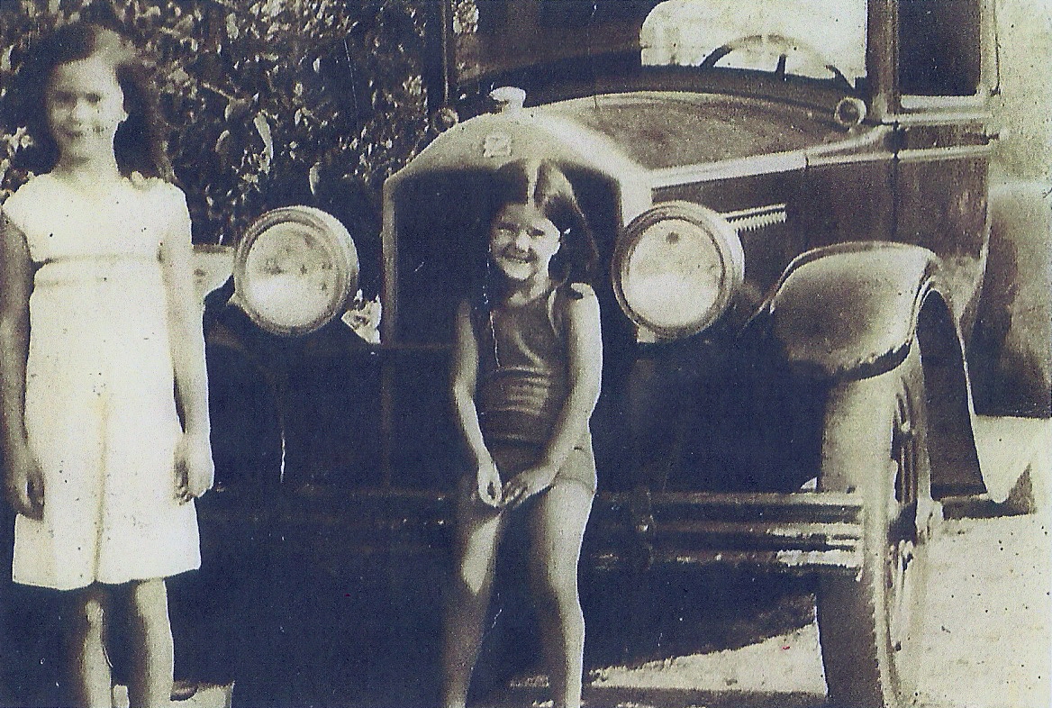 A black and white photo of two young girls, Bette Benavides and her sister Ange pose in front of their family car in 1937. One girl stands and the other sits on the front bumper.
