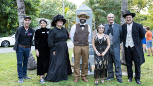 A group of people dressed in historical costumes stand together in front of a tall headstone inside of a cemetery. Green grass and trees can be seen in the background.