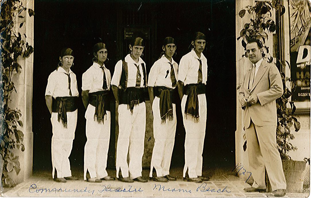 A black and white photo from 1927 with 5 men standing in a row facing the camera, wearing white outfits with a waist sash, head scarf, and hoop earrings. They are "community theater ushers." A man in a suit stands apart from the men looking at the camera.
