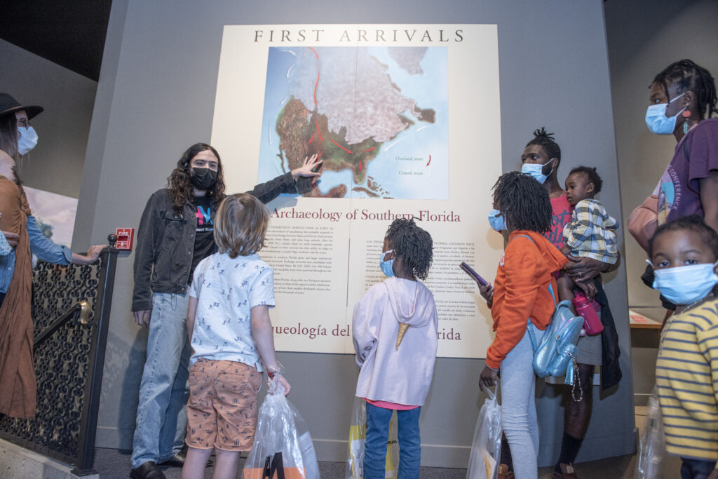 A museum educator stands in front of a group of people in a museum exhibition. They point to a map on the wall.