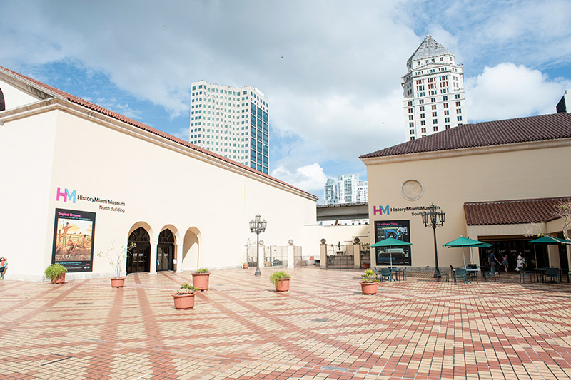 View of the Miami-Dade Cultural Plaza, with yellowy-beige and orange tiles, with HistoryMiami Museum's two buildings in view. In the background is the Miami-Dade County Courthouse against a blue sky.