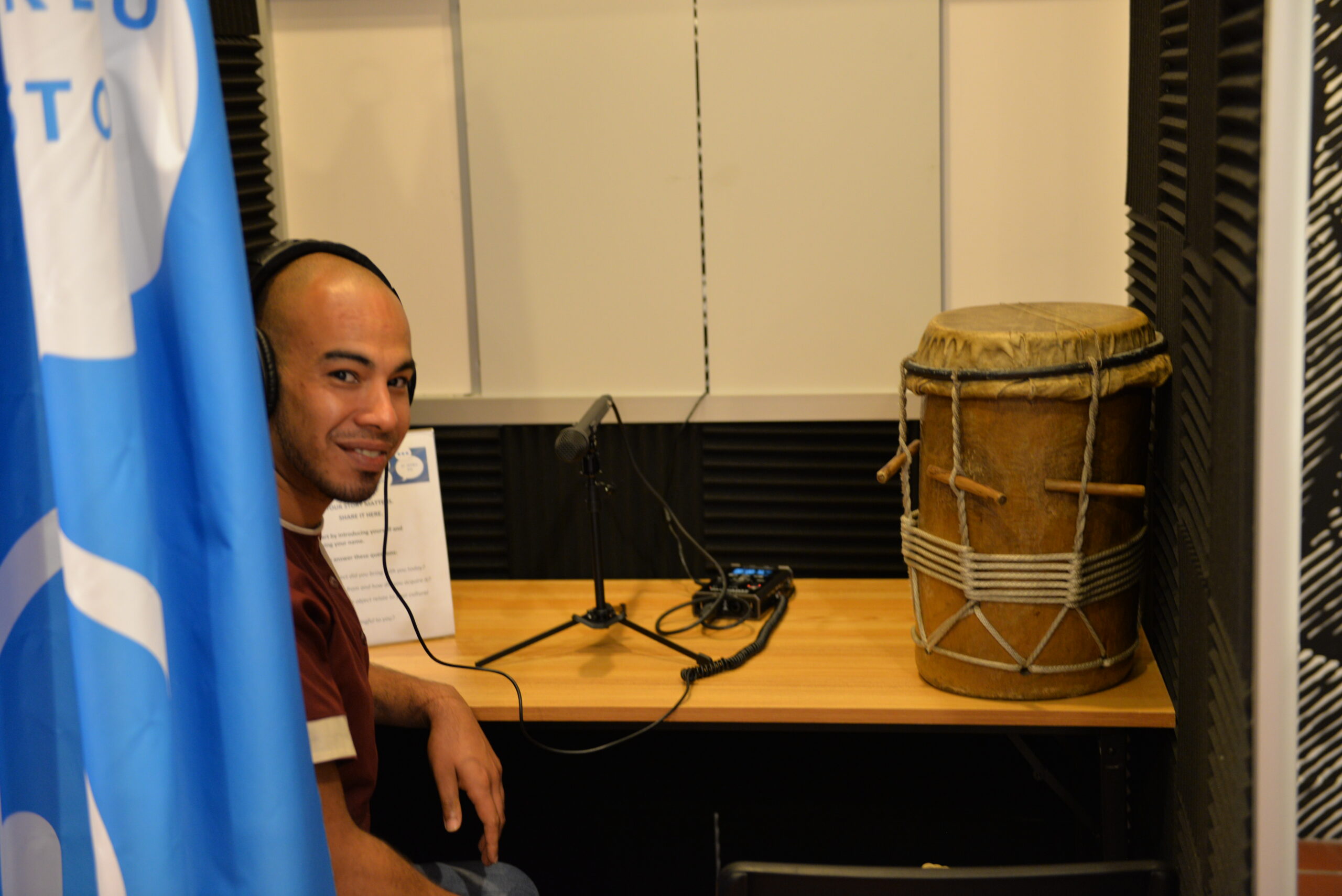 a man is sitting inside of a booth with headphones on and a microphone in front of him on the table. A drum made of wood sits on the table next to him.