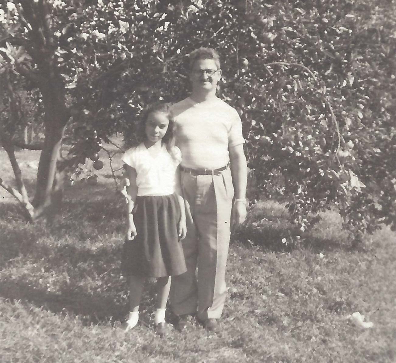 A black and white photo of a young girl, Diane Bronstein, and a man, James Betesh, stand in the grass of an orange grove in 1947