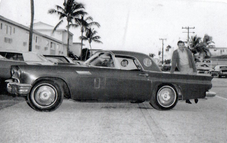 A black and white photo of a man, Ed Herder, standing near the trunk of his car, a 57 Ford T Bird