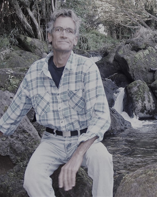 A man sits on top of a rock with trees and a stream in the background.
