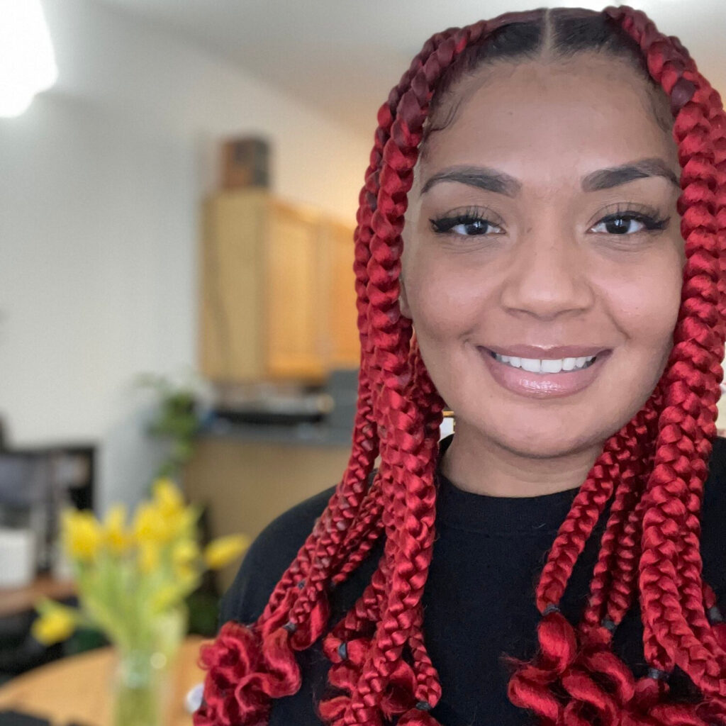 A person wearing a black top and red multi-braided hair smiles towards the camera.
