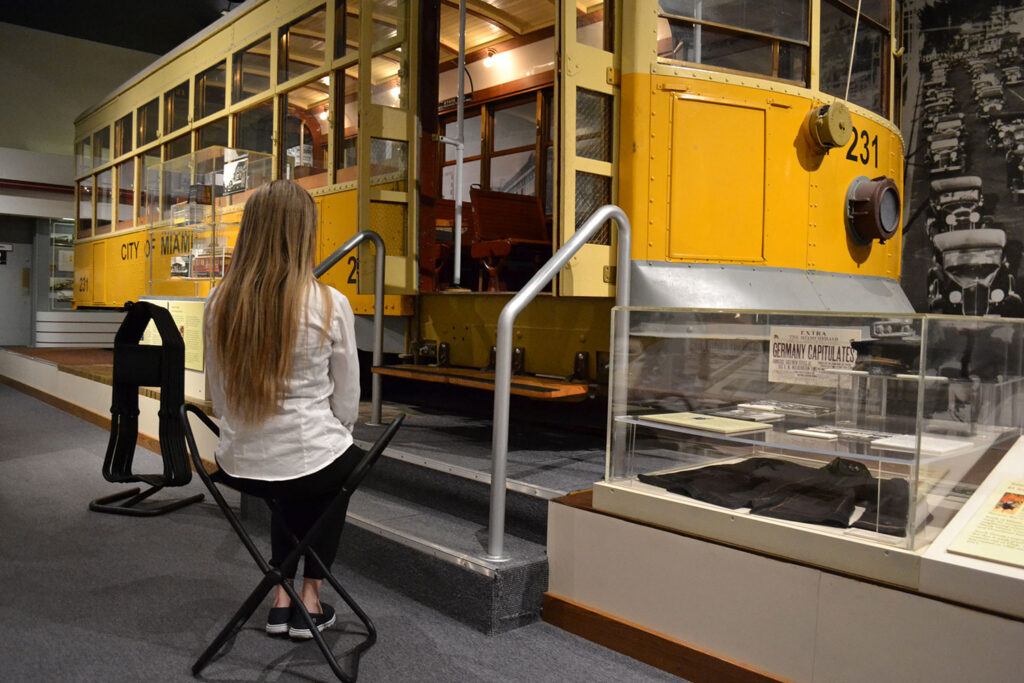 A person wearing a white top, black pants, and black sneakers sits on a portable stool in front of the steps towards a large yellow trolley inside of a museum exhibition.