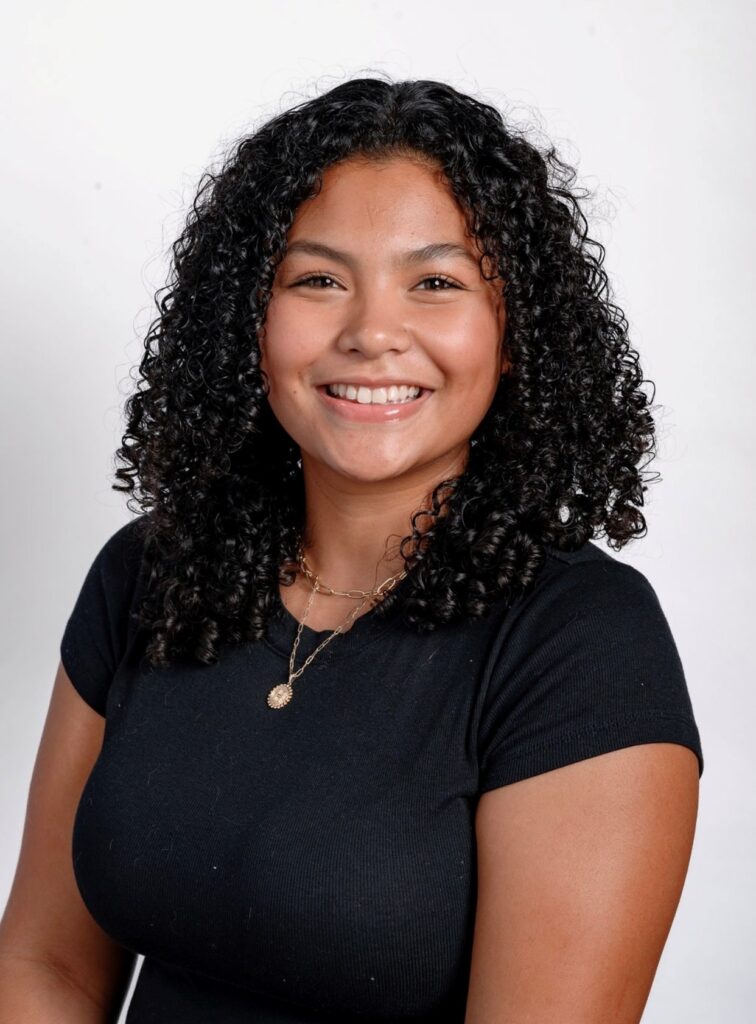 A person with shoulder-length curly dark brown hair wearing a black shirt smiles towards the camera.
