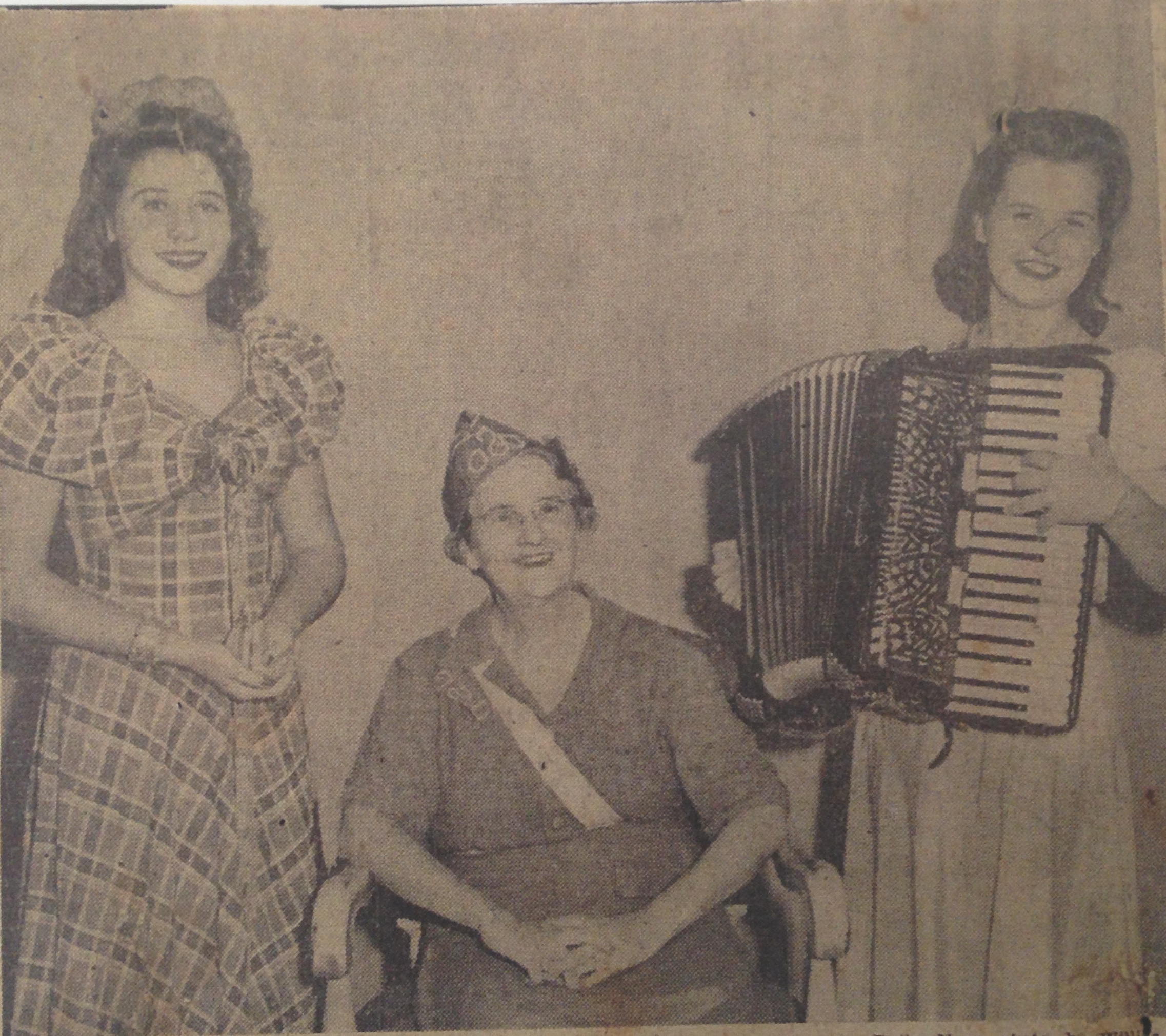 A black and white photo three women. Two younger women are standing, one is holding an accordion. An older woman is sitting in a chair between them.