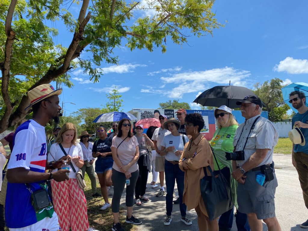 A tour guide wearing a straw hat and blue and white Haitian shirt stands in front of a group of tour participants in Little Haiti on a sunny day.
