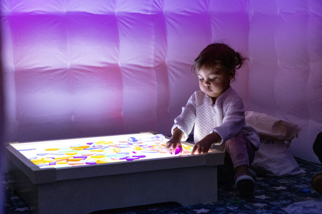 Child in a purple-lit room plays with colorful pebbles on a light table