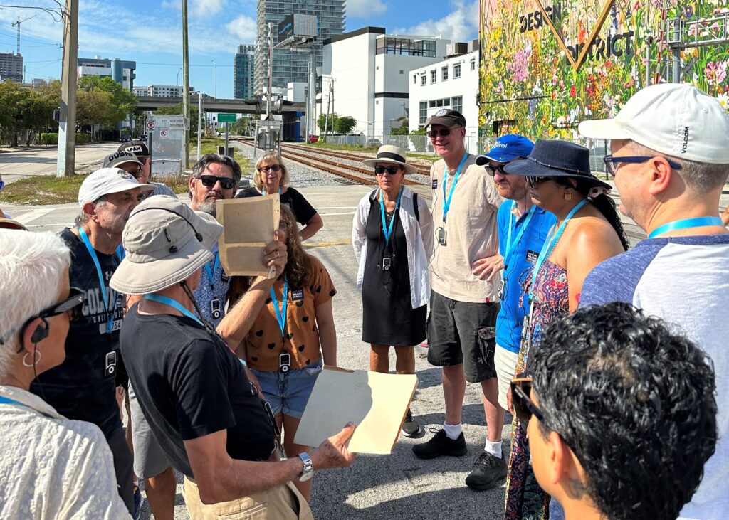 A group stands on the side of a paved road in the Design District. They look towards a tour guide who stands wearing a hat, black shirt, and shorts and is holding a piece of paper up to the group.