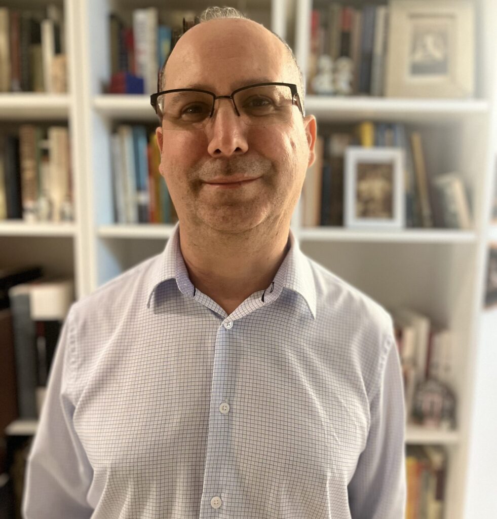 A person with short, shaved hair stands in front of a bookshelf. They wear half dark rimmed glasses and a pastel checked button down shirt as they smile towards the camera.